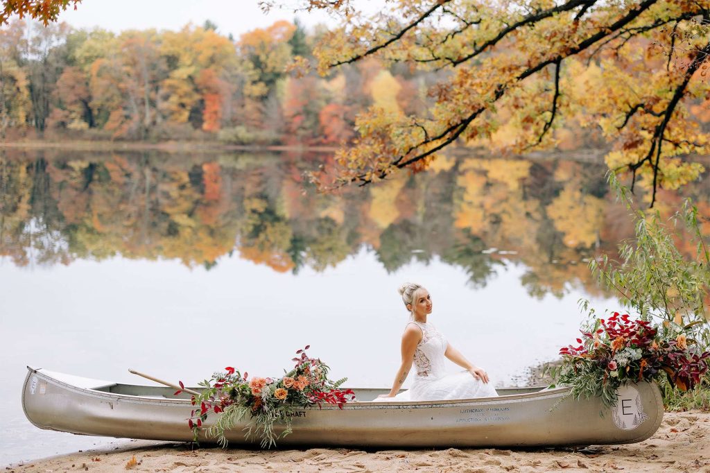 Bride sits in floral drenched canoe on a mirrored lake at Camp Elsewhere in Waupaca, WI .