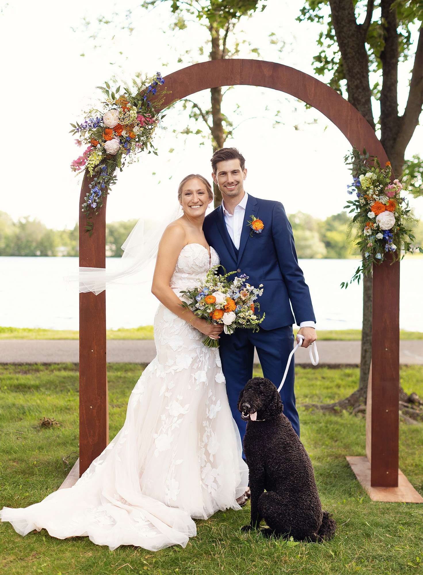 Bride, groom, and dog pose under arch adorned with bright hued florals.