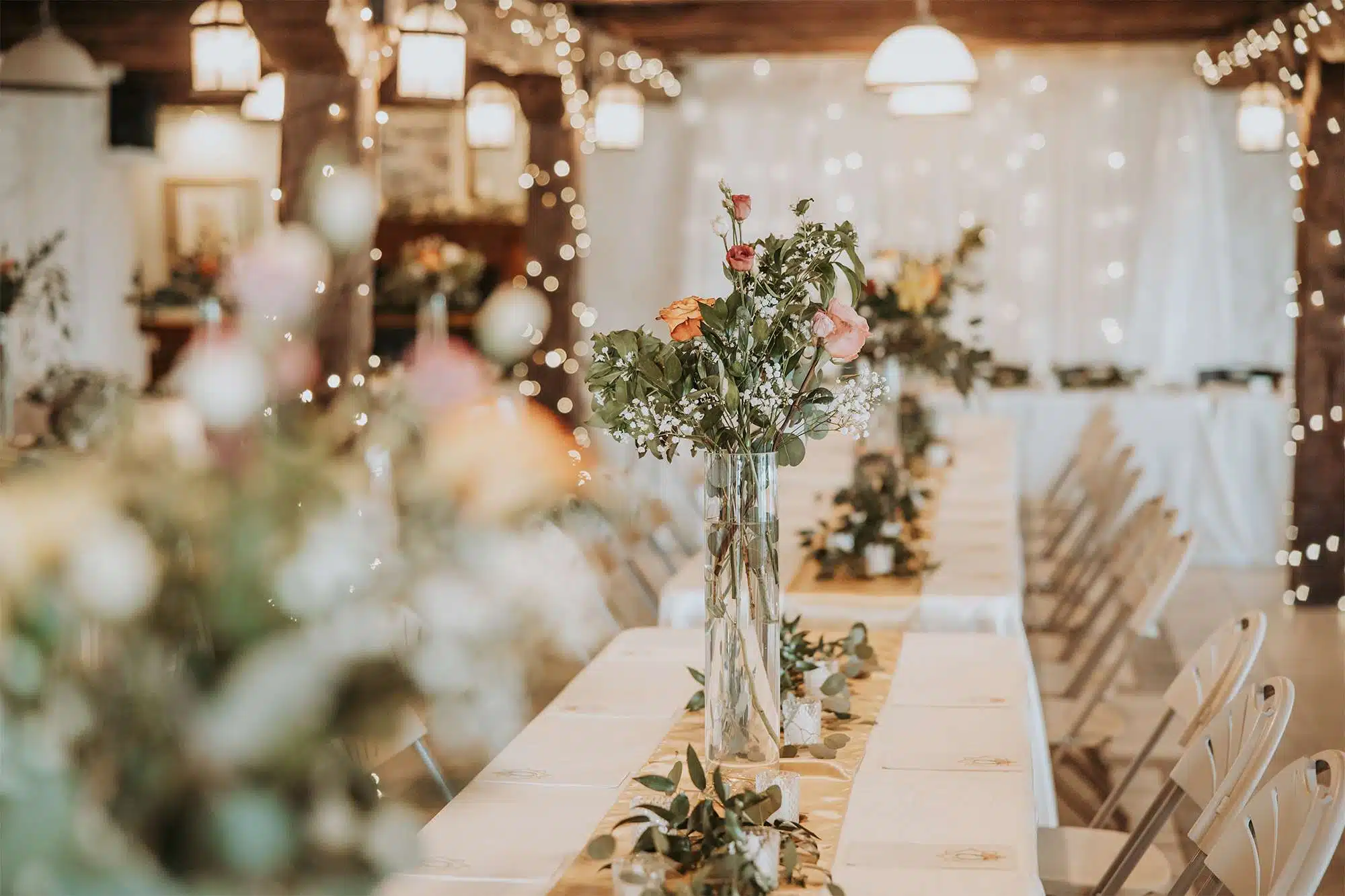 Wedding reception table with tall floral centerpieces and greenery by Archer & Bliss Floral, set in a softly lit rustic indoor venue.