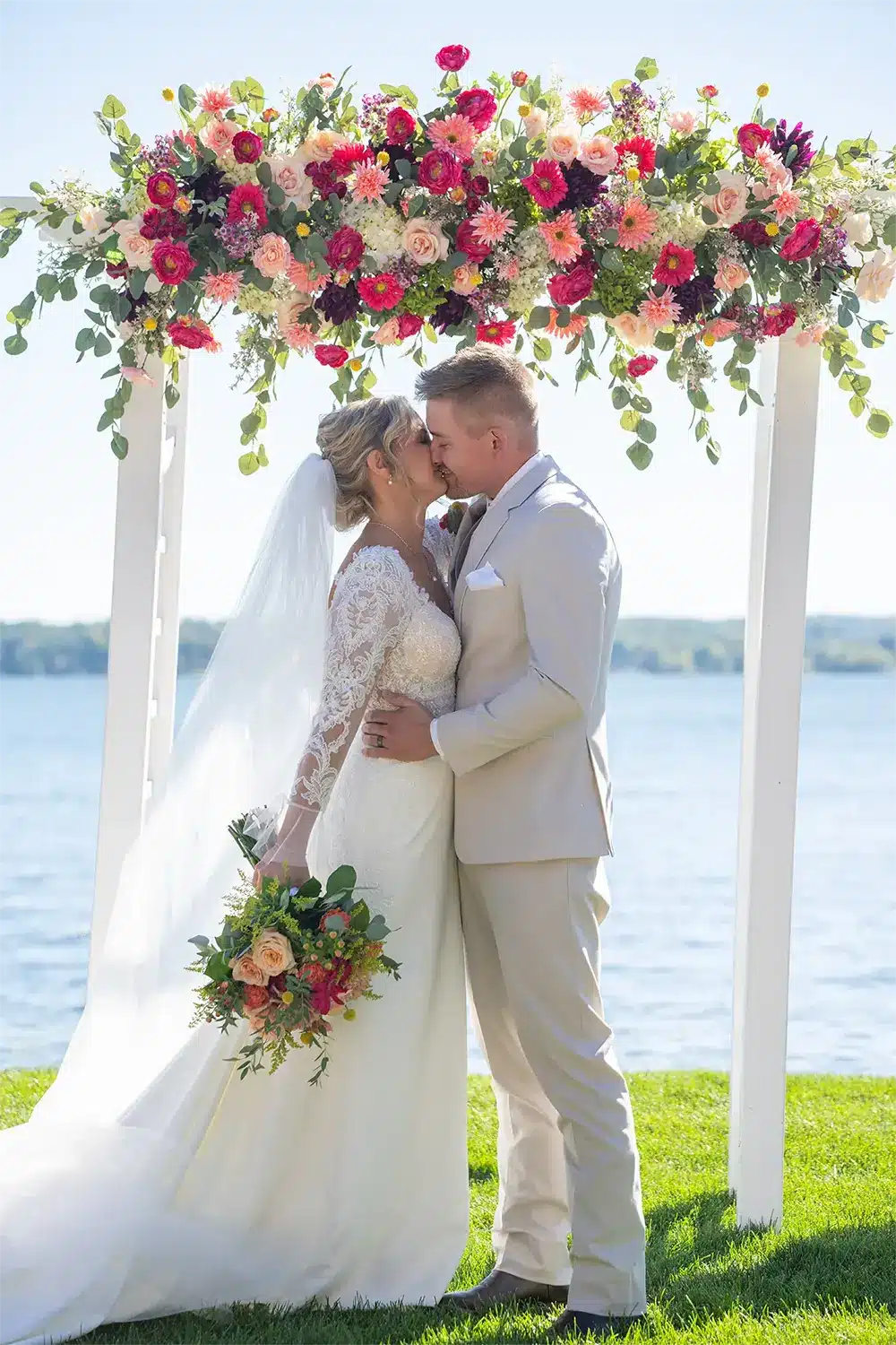 Bride and groom kiss under floral drenched white arch created by I Do Decorating . Bride and groom kiss under floral drenched white arch created by I Do Decorating .