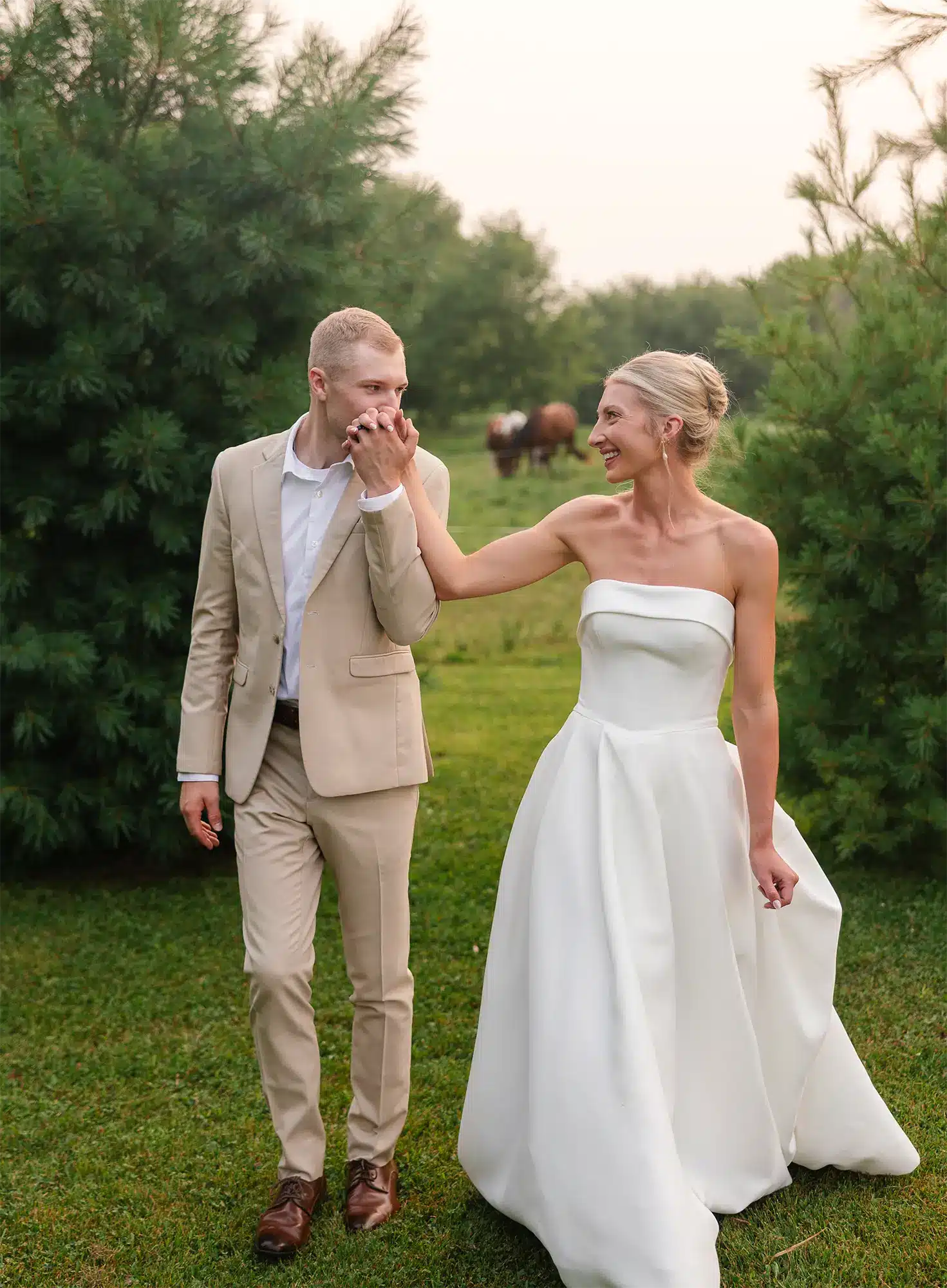 Faith-Filled Summer Wedding in Cecil, WI- Bride and groom Bride and groom walking hand in hand in a grassy field, groom kissing bride’s hand with horses in the background