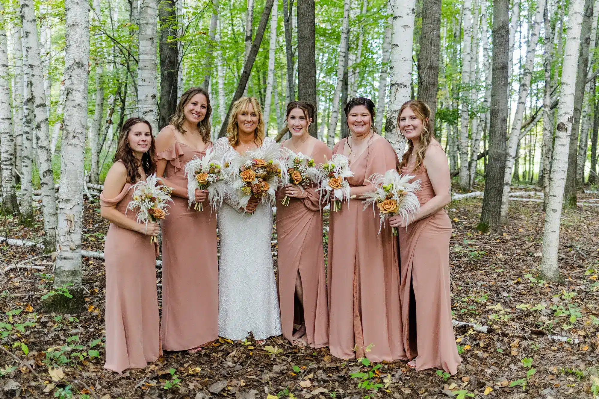 Bride with bridesmaids in blush dresses holding boho-style bouquets in a wooded setting.