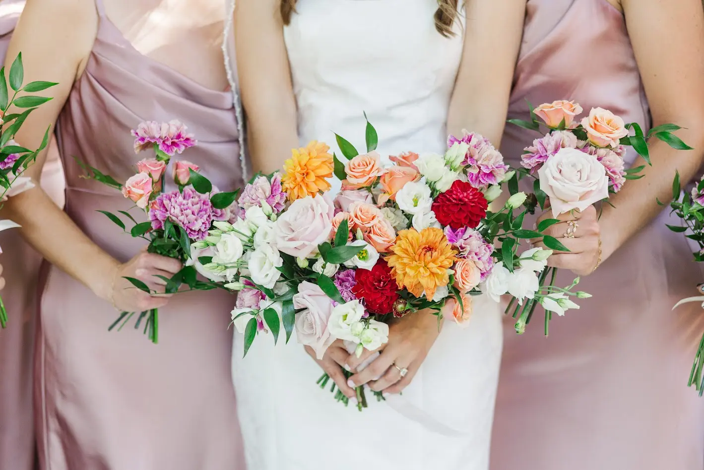 Bride and bridesmaids holding colorful bouquets with roses, dahlias, and greenery.