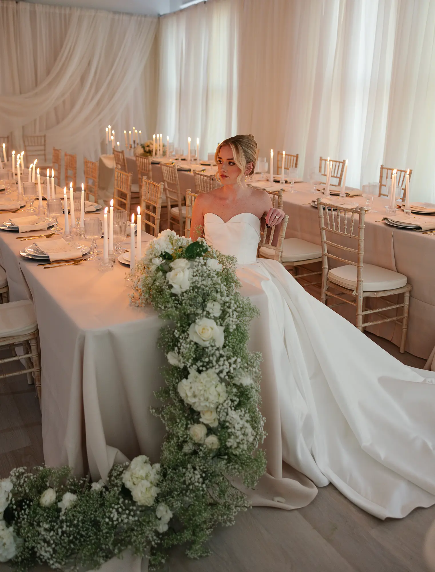 Elegant Candlelit Wedding Reception with Cascading Florals. Bride seated at candlelit reception table with cascading white floral arrangement and greenery.