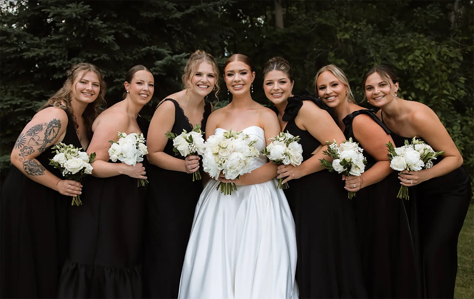 Bride with bridesmaids in black dresses holding white rose bouquets outdoors.