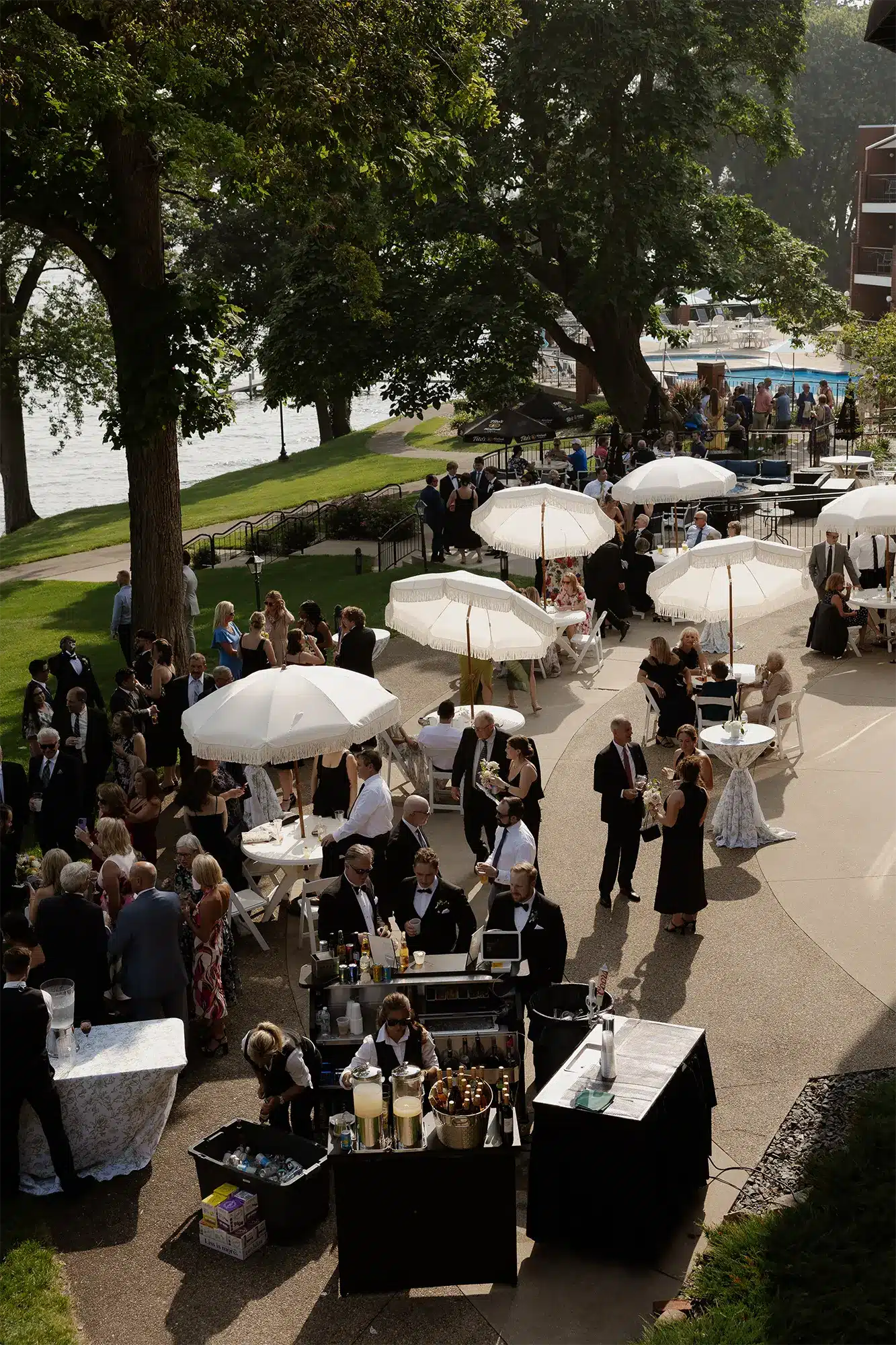 Outdoor Cocktail Hour at Heidel House Wedding on Green Lake Aerial view of wedding cocktail hour on the Heidel House patio with guests mingling under white fringed umbrellas, mobile bar service, and Green Lake visible through the trees.