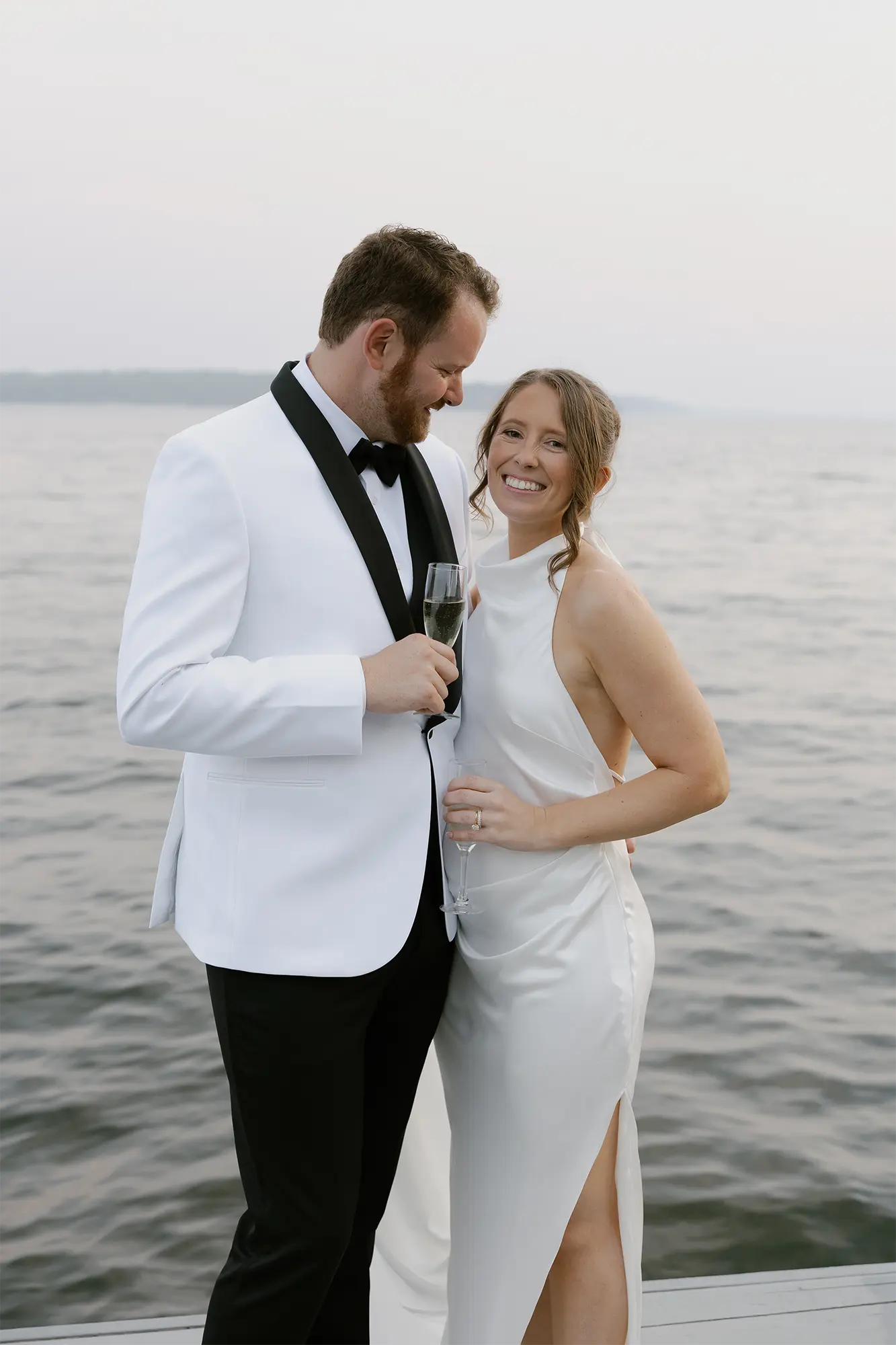 Elegant Waterfront Portrait at Heidel House | Green Lake Wedding Bride and groom in modern white attire toasting with champagne on dock at Heidel House Green Lake waterfront wedding.