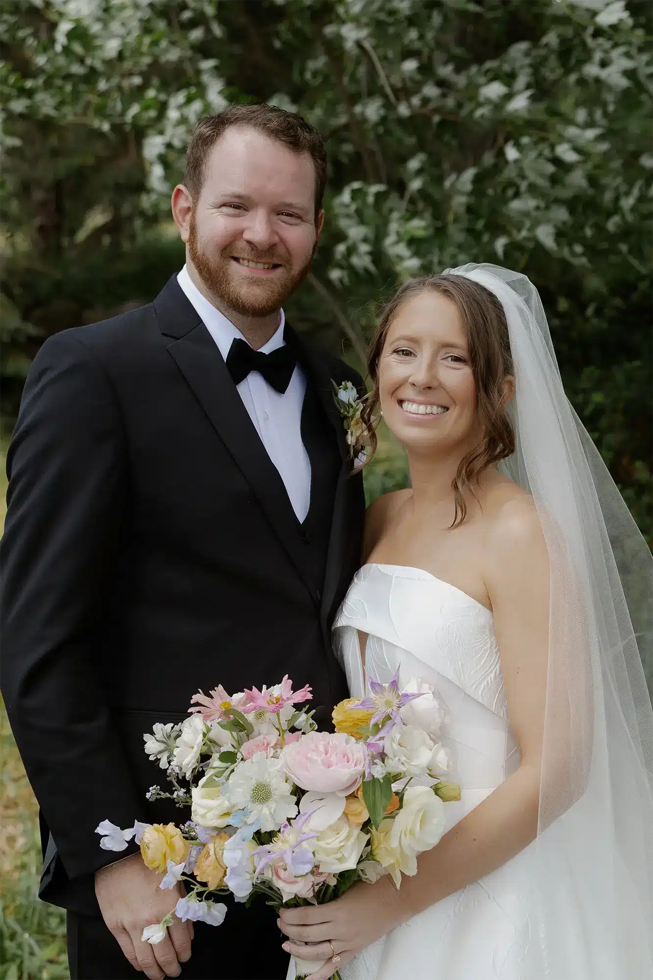 Caitlin and Charles Wedding Portrait at Heidel House on Green Lake Bride in white strapless gown and veil holding a pastel wildflower bouquet standing with groom in black tuxedo and bow tie in front of green garden foliage at the Heidel House.
