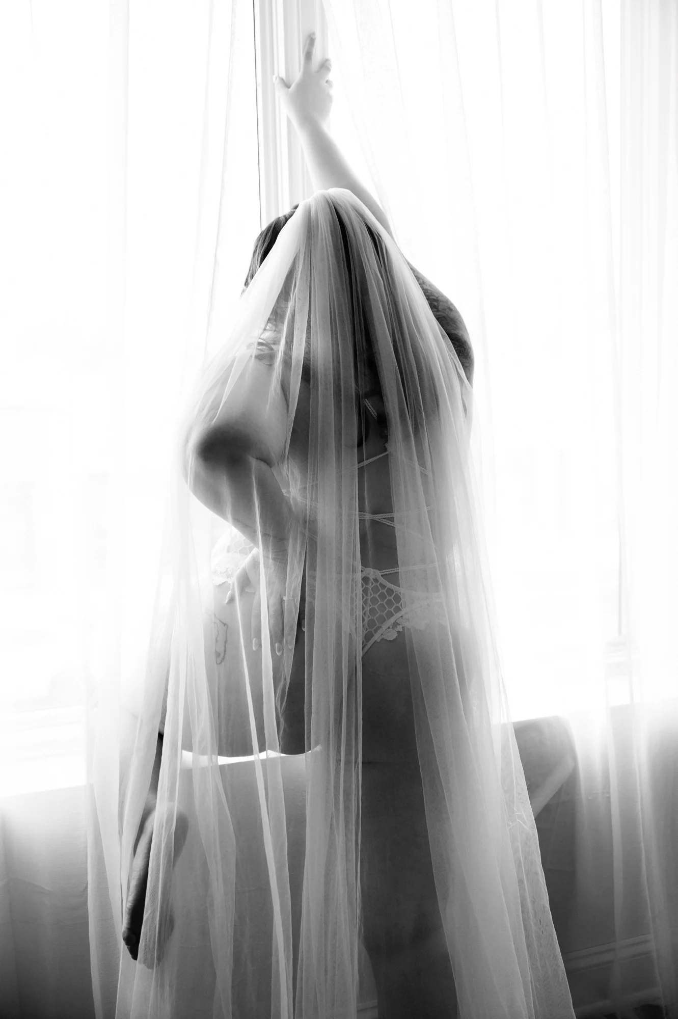 Woman wrapped in a bridal veil standing in front of a bright window.