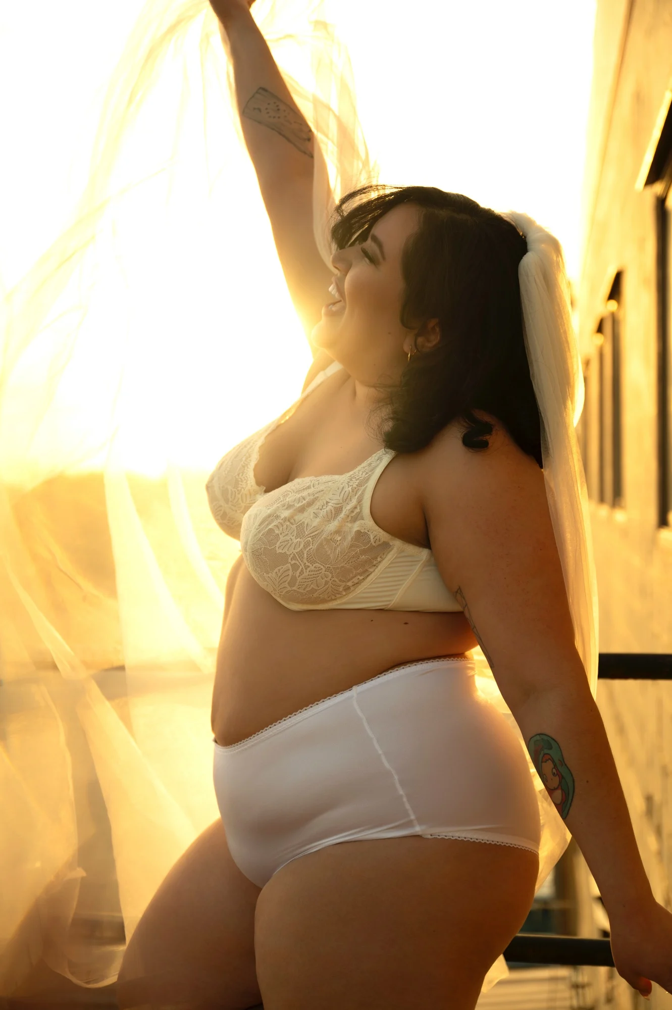 Woman in white lingerie standing in warm sunlight beside flowing curtains.